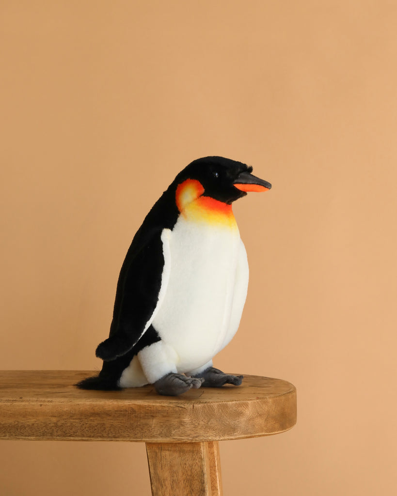 The Penguin Stuffed Animal, Small, features black, white, and orange colors with realistic details. It is displayed sitting on a wooden bench against a beige background, standing out among other plush toys.