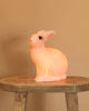 The White bunny Lamp, with its smooth white surface and adorable features, sits on a rustic wooden stool against a beige background, casting a soft, warm glow as a whimsical night light for any nursery.