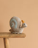 The Steiff Snaily Snail Stuffed Plush Animal, 8 inches, with a light gray body, yellow antennae, and blue spiral shell, sits on a wooden stool against a plain beige background.