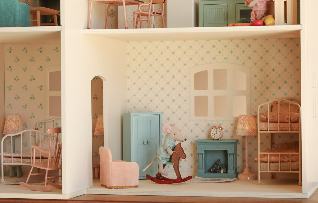 The Maileg Fully Furnished Dollhouse Set + Surprise Gift features a charming dollhouse interior complete with pastel-colored furniture, patterned wallpaper, cozy bunk beds, a classic rocking chair, and a toy mouse riding a wooden rocking horse as the centerpiece, bringing the enchanting world of miniatures to life.