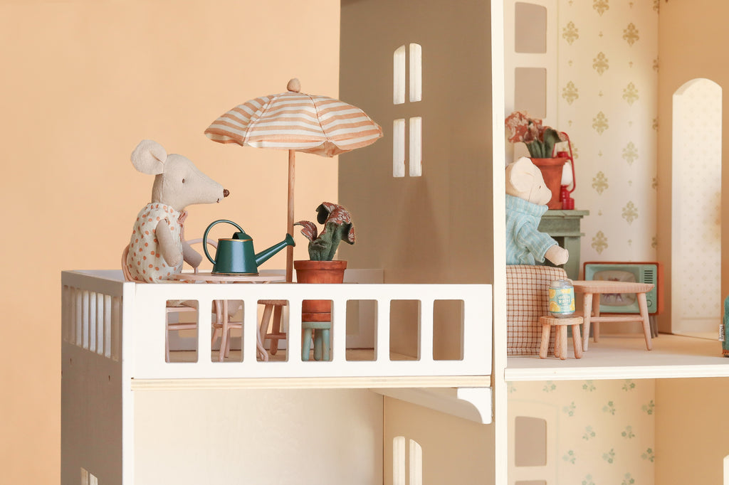 Stuffed toy mice are arranged in the Maileg Fully Furnished Dollhouse Set + Surprise Gift, with one perched on a balcony holding a watering can and tending to a potted plant under a striped umbrella, while another relaxes inside near miniature toy furniture.