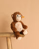 The Bodo Monkey Plush Stuffed Toy, 12 Inches, in brown and white with a stitched smile, sits on a wooden bench against a beige background. A yellow tag in its ear marks it as an authentic Steiff product.