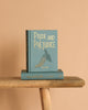 The "Pride and Prejudice | Austen | Collector's Ed. | Hardcover," featuring a teal cover with a gold peacock design, stands upright on another matching copy, both placed on a rustic wooden bench against a beige background.
