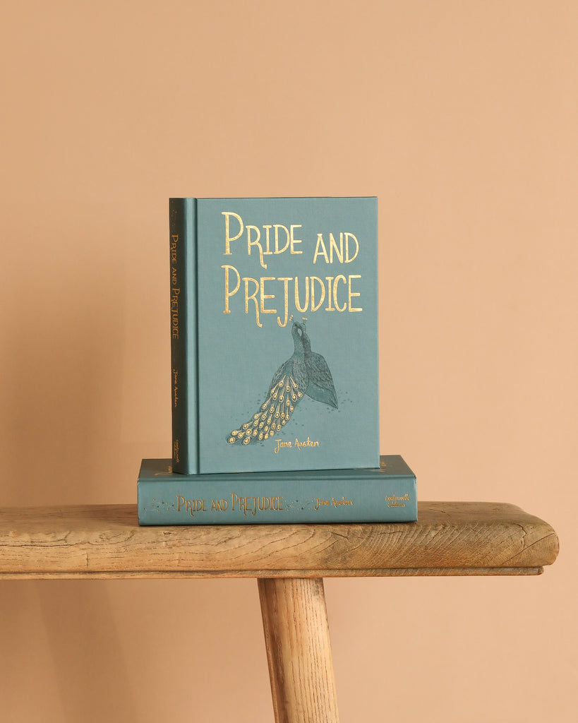 The "Pride and Prejudice | Austen | Collector's Ed. | Hardcover," featuring a teal cover with a gold peacock design, stands upright on another matching copy, both placed on a rustic wooden bench against a beige background.