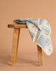 An Organic Cotton Patchwork Baby Blanket - Spring Bloom, with blue plaid lines, embroidered flowers, and scalloped edges, is draped on a wooden stool against a plain beige background.