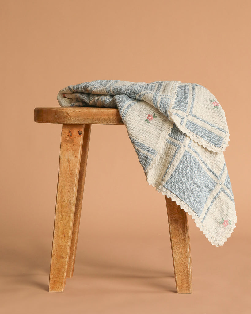 An Organic Cotton Patchwork Baby Blanket - Spring Bloom, with blue plaid lines, embroidered flowers, and scalloped edges, is draped on a wooden stool against a plain beige background.