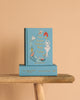 Two blue hardcover copies of "Hans Christian Andersen Fairy Tales | Collector's Edition" with whimsical illustrated covers are stacked on a wooden bench—one upright, one flat—against a light beige background.