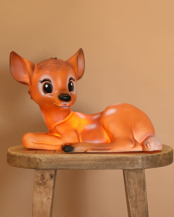 The Resin Deer Lamp features a cartoon-style fawn with big eyes lying on its side atop a wooden stool, creating a delightful decor accent for children's rooms, shown against a plain light brown background.