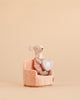 A Maileg Little Sister with Tooth Box Set mouse, dressed in pink, sits on a Maileg Soft Folding Chair and holds a purple heart-shaped tooth pillow against a plain beige background.