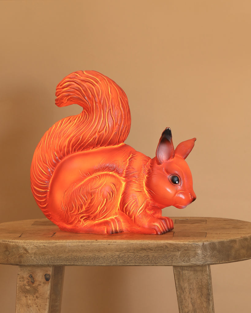 The Squirrel Lamp is a bright orange figurine with a bushy tail, perched on a wooden stool against a beige background—perfect as a playful night light or charming decor for any nursery or child's room.