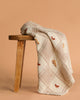 The Organic Cotton Baby Blanket - Farm, featuring a cream hue, textured checkered pattern, crochet trim, and embroidered birds, is draped over a wooden bench against a solid beige background.
