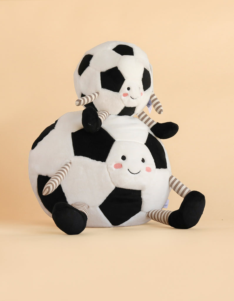 Two plush toys from the Mon Ami Soccer Ball Set, each shaped like a soccer ball with smiling faces and striped arms and legs, sit together on a beige background.