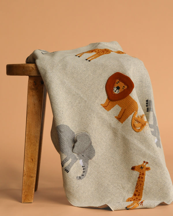 The Organic Cotton Knit Baby Blanket - Jungle, in light gray with cartoon lion, elephant, and giraffe designs, is draped over a wooden stool against a plain beige background.
