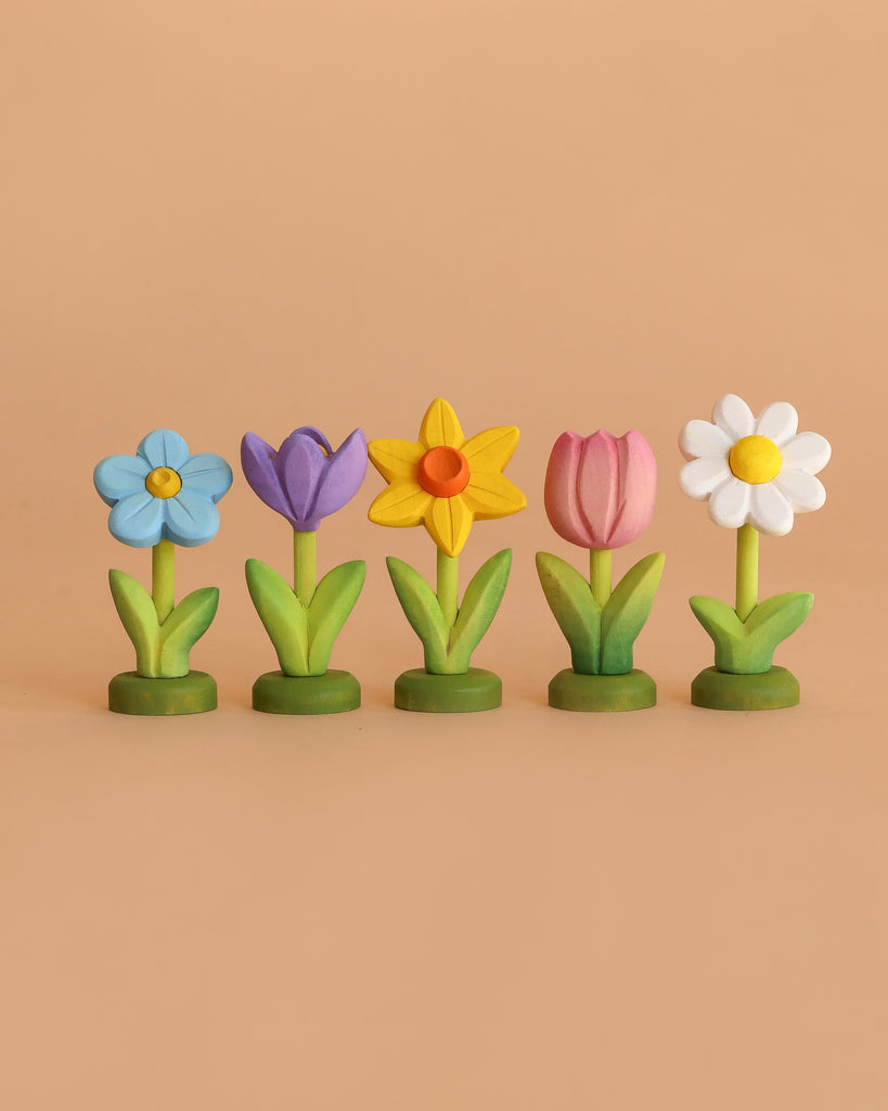 The Bumbu Handmade Wooden Celebration Ring features five upright wooden flowers in blue, purple, yellow, pink, and white on green bases—eco-friendly toys that inspire creative Montessori play against a simple beige background.