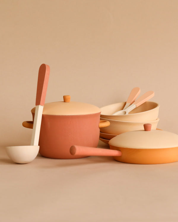 A Handmade Wooden Kitchen Essentials - Flower set in muted earth tones, including a large pot with a lid, a frying pan, a ladle, and bowls with spoons, all arranged neatly against a beige background. The items have a minimalist design and complementary colors in shades of beige and terracotta.