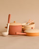 A Handmade Wooden Kitchen Essentials - Flower set in muted earth tones, including a large pot with a lid, a frying pan, a ladle, and bowls with spoons, all arranged neatly against a beige background. The items have a minimalist design and complementary colors in shades of beige and terracotta.
