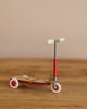 The Maileg Mouse Size Scooter / Kickboard - Red, with white wheels and wooden details, stands on a wooden surface. It features a sleek metal stick with small deck and handlebars, set against a plain beige wall.