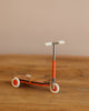 The Maileg Mouse Size Scooter in orange rests on wood, showing off its wooden details against a plain beige background. It features a T-shaped handlebar and white wheels, making it perfect for tiny riders.