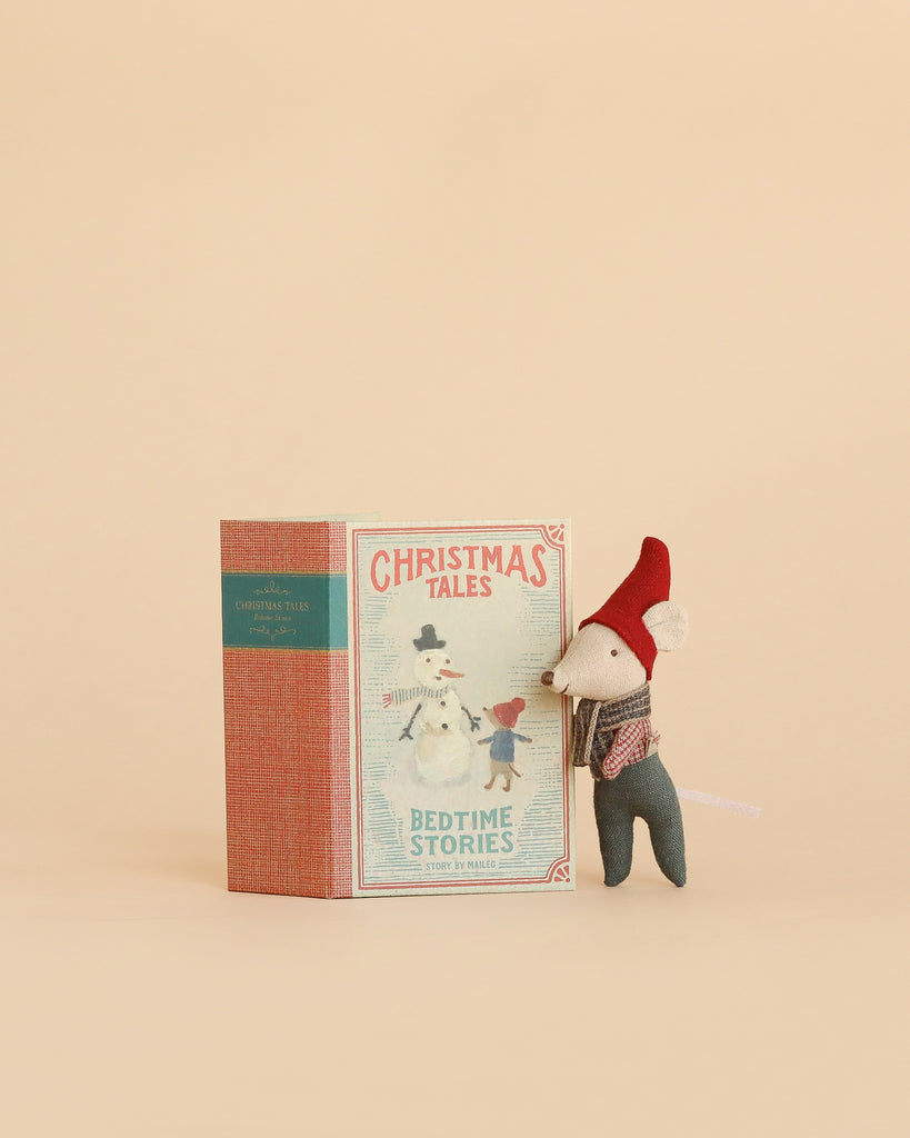 A delightful Maileg Christmas Mouse, Baby in Book, wearing a red hat, stands next to the “Christmas Tales: Bedtime Stories” illustrated book featuring two sweet mice on the cover, all set against a simple beige background.