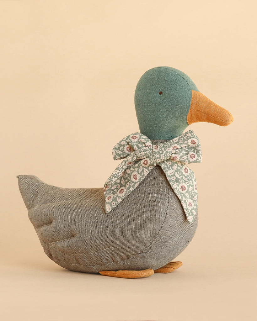 The Maileg Duck with Bow - Grey features a plush design with a teal head, orange beak and feet, soft gray body, and a charming floral-patterned bow around its neck. Set against a beige background, it makes an ideal Christmas duck decoration, adding an authentic festive touch to your seasonal animals display.