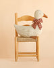 The Maileg Duck with Bow - Sand, featuring a red patterned bow and soft plush design, sits on a wooden chair with a woven seat against a light beige background—perfect as a charming Christmas duck decoration.