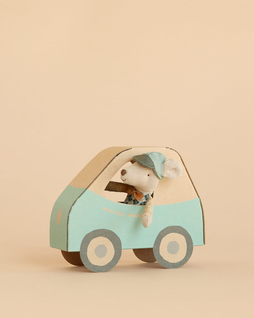 A Maileg Driver Mouse, Little Brother, wearing a blue cap looks out the window of a toy mouse car crafted from cardboard and painted in blue and beige, set against a plain beige background.