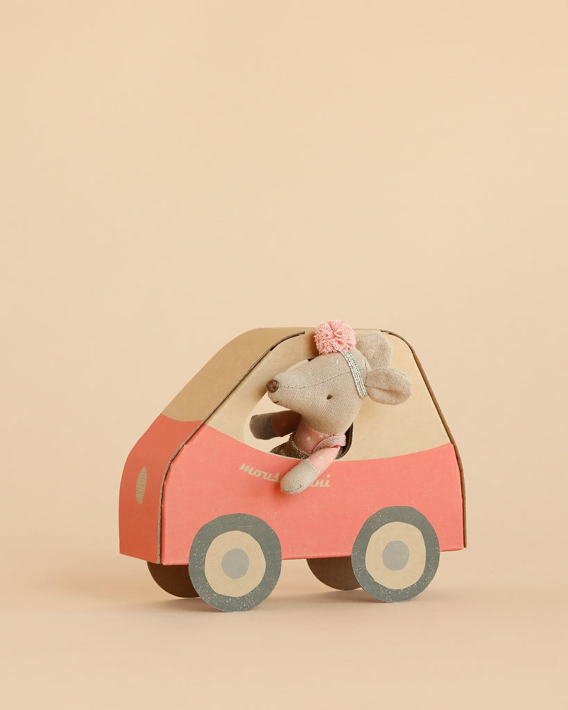The Maileg Driver Mouse, Little Sister, wearing a headband with a pink flower, sits inside a red and beige mouse car with round gray wheels against a plain beige background.