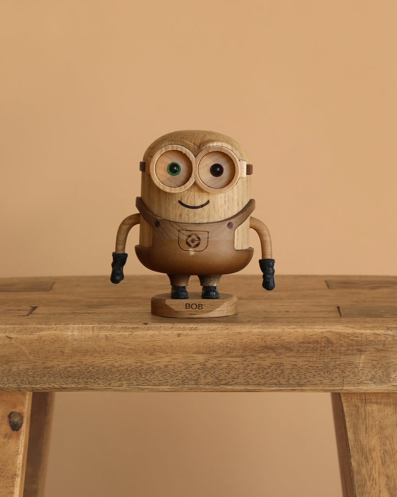The Boyhood Minion Bob wooden figurine, featuring mismatched eyes and a "BOB" label on its base, stands on a wooden table with a plain beige background.