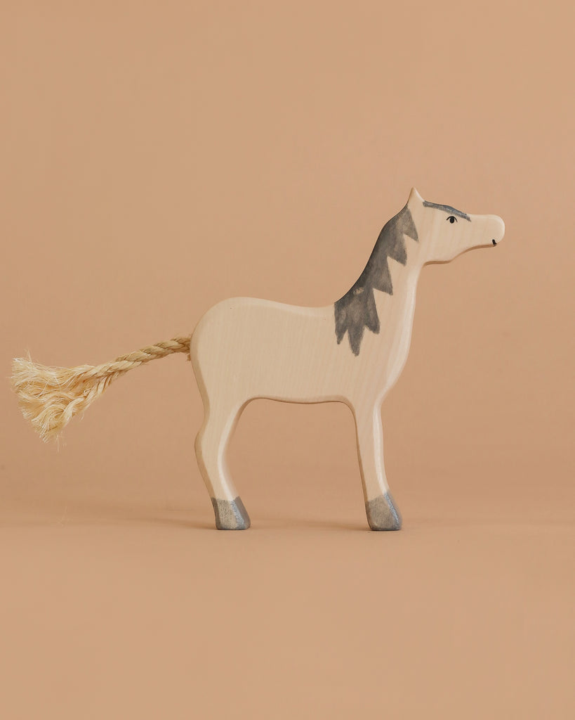 The Holztiger Grey Horse, Head Raised is a handcrafted wooden toy with simple painted details, gray mane, and rope tail, standing on a beige surface—a charming example of European craftsmanship.