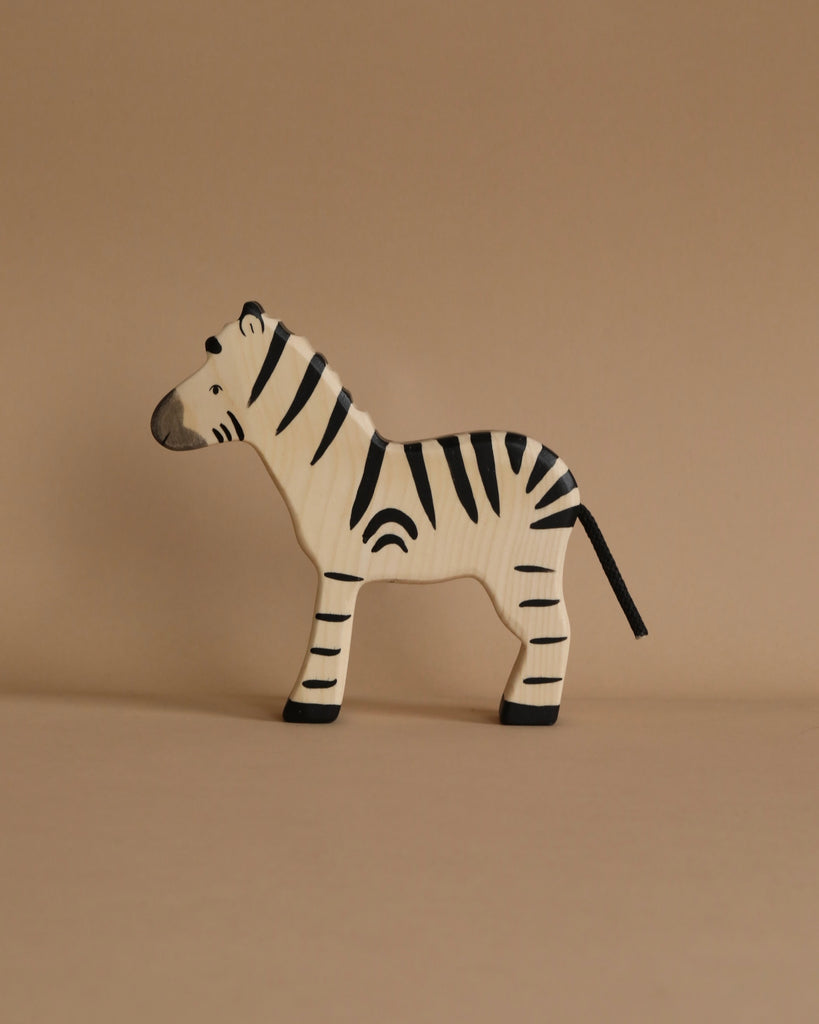 A small, handmade zebra from the Holztiger Handmade Wooden Safari Animals Set with black stripes stands on a beige surface and background—ideal for collectors of animal figurines and fans of Holztiger figures.