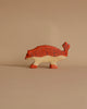 A small, hand-carved wooden toy in the shape of an animal, painted in shades of orange and brown with patterned markings along its body. This Holztiger Ankylosaurus Dinosaur has four legs, a tail lifted upwards, and a pointed snout, reminiscent of HOLZTIGER figures, set against a plain beige background.