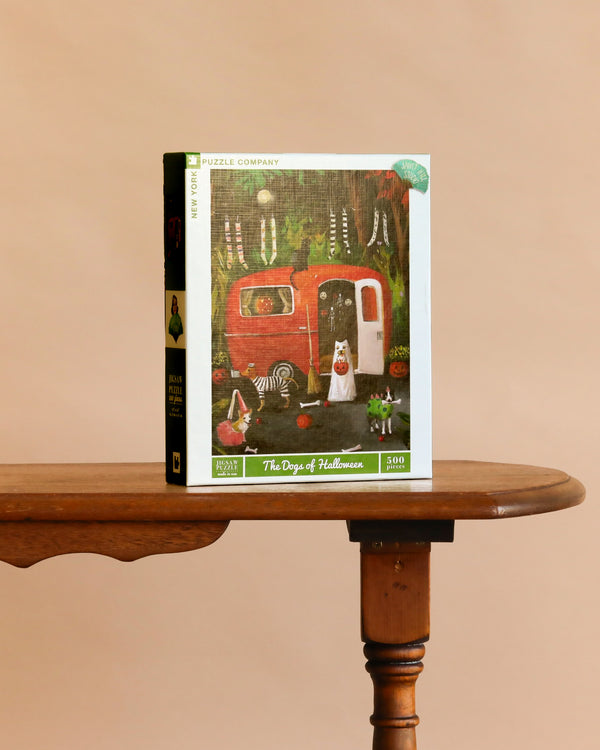 The Dogs of Halloween - 500 Piece Jigsaw Puzzle sits on a wooden table, featuring an illustration of costumed dogs near a red trailer amid Halloween decorations.