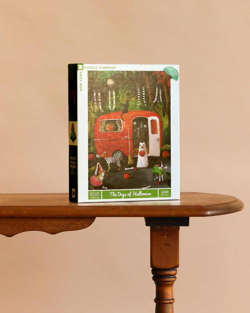 The Dogs of Halloween - 500 Piece Jigsaw Puzzle sits on a wooden table, featuring an illustration of costumed dogs near a red trailer amid Halloween decorations.