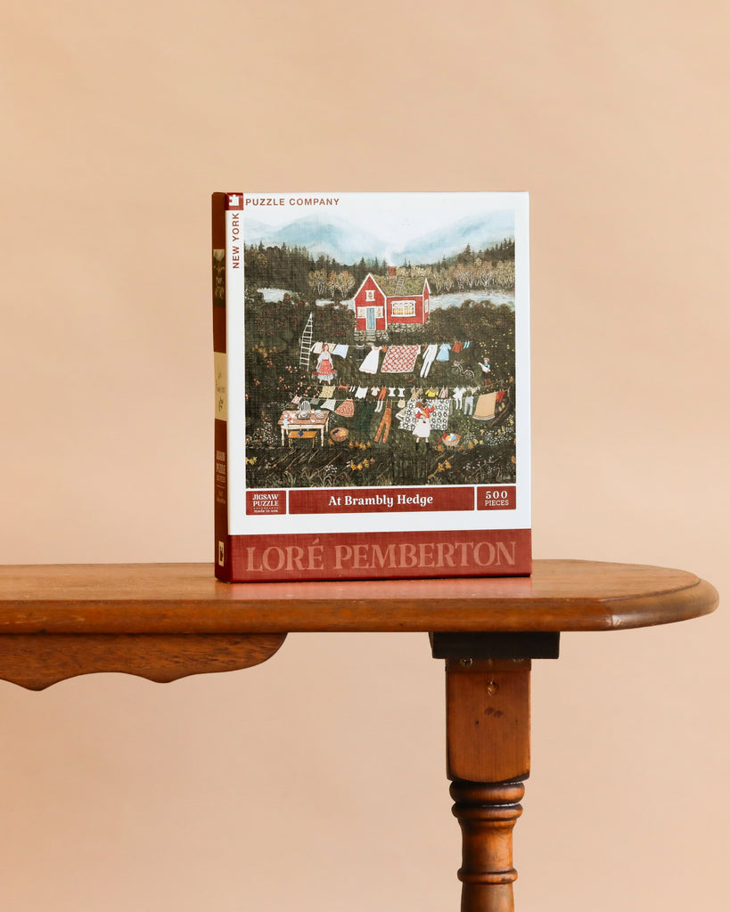 At Brambly Hedge - 500 Piece Jigsaw Puzzle features a country scene with gardens, laundry on a line, and a red house. Displayed upright on a wooden table against a beige background. Made in USA.