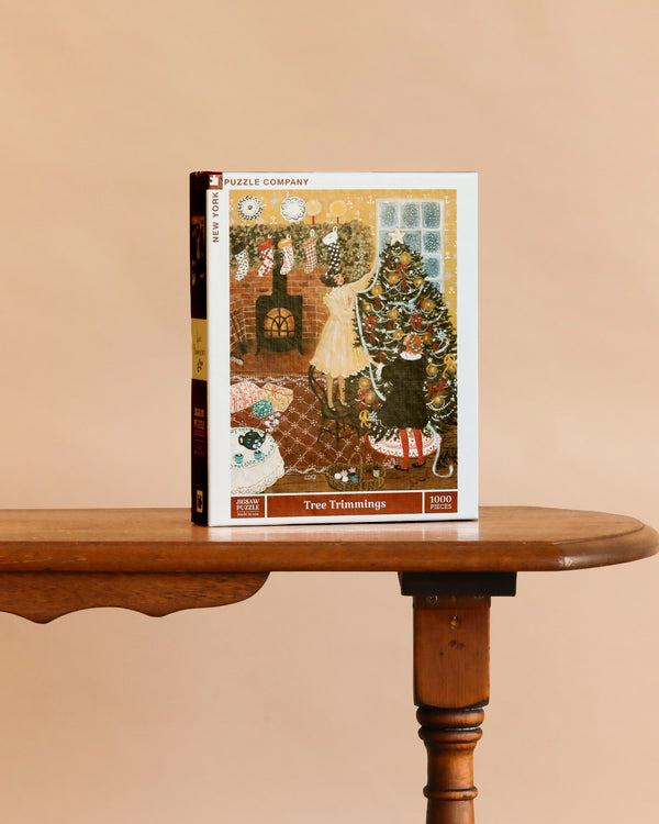 A “Tree Trimmings - 1000 Piece Jigsaw Puzzle” box stands upright on a wooden table, showing an illustration of a woman decorating a Christmas tree by the fireplace.