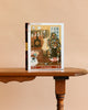 A “Tree Trimmings - 1000 Piece Jigsaw Puzzle” box stands upright on a wooden table, showing an illustration of a woman decorating a Christmas tree by the fireplace.