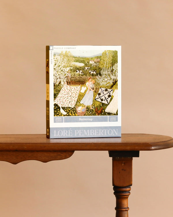 A box of Buttercup - 500 Piece Jigsaw Puzzle sits upright on a wooden table, its linen-style finish showcasing women with quilts in a scenic pastoral landscape.
