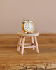 A small yellow alarm clock sits on a light wooden stool, both positioned on a rustic wooden surface with a neutral background—the perfect setting to showcase the Maileg Family Room Set as delightful miniature décor or charming dollhouse accessories.