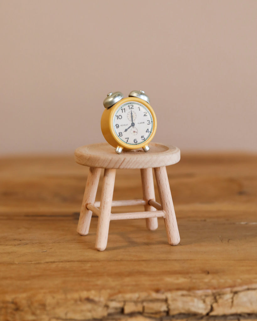 A small yellow alarm clock sits on a light wooden stool, both positioned on a rustic wooden surface with a neutral background—the perfect setting to showcase the Maileg Family Room Set as delightful miniature décor or charming dollhouse accessories.