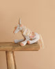 The Maileg Unicorn with Suitcase Set features a soft, light gray unicorn with a pastel mane and tail, sitting on a rustic bench—perfect for collectors or pairing with other Maileg accessories.