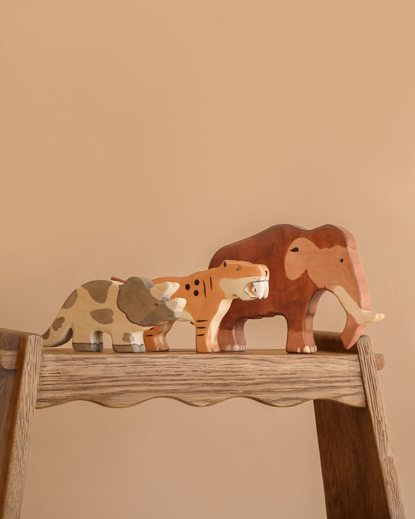 The Holztiger Handmade Wooden Prehistoric Animals Set features a triceratops, saber-toothed cat, and mammoth, beautifully displayed on a wooden surface against a plain beige background.