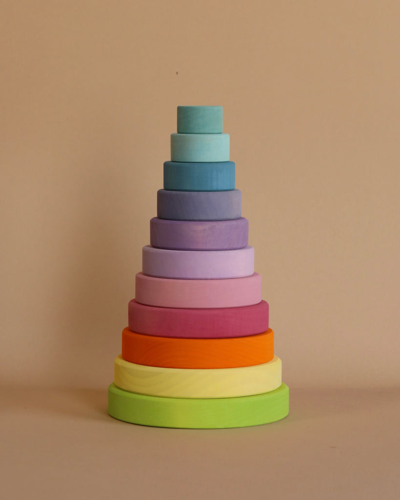 A neatly stacked pyramid of colorful wooden rings arranged by size from largest to smallest, with the largest ring at the bottom in green and the smallest at the top in light blue, all set against a plain beige background. This Pastel Stacking Tower enhances fine motor skills and eye-hand coordination.