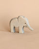The Ostheimer Handmade Wooden Safari Animals Set features a small, light gray handcrafted wooden elephant with simple carved details, standing on a beige surface—perfect for imaginative play.