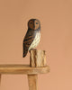 The Hand Carved Ornament - Barred Owl - 13"H features brown and white markings and is perched on a small tree stump, displayed on a wooden stool with a plain light brown background.