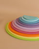 A set of colorful, semi-circular wooden stacking toys, reminiscent of modern architecture, is arranged in a gradient from largest to smallest. The pieces are in various shades of green, yellow, orange, red, pink, purple, and blue. The background is a plain beige surface.