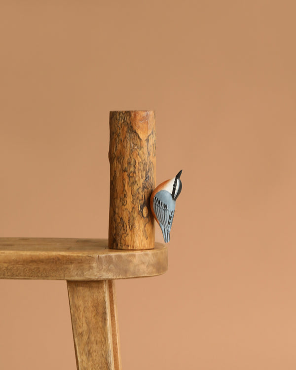 The Hand Carved Ornament - Red Breasted Nuthatch Bird- 8"H is shown clinging to a sustainable softwood tree log, displayed on a wooden table with a plain brown background.