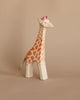The Ostheimer Handmade Wooden Safari Animals Set features a handcrafted wooden giraffe with orange spots and a pink detail on its head, standing against a beige background—perfect for imaginative play.