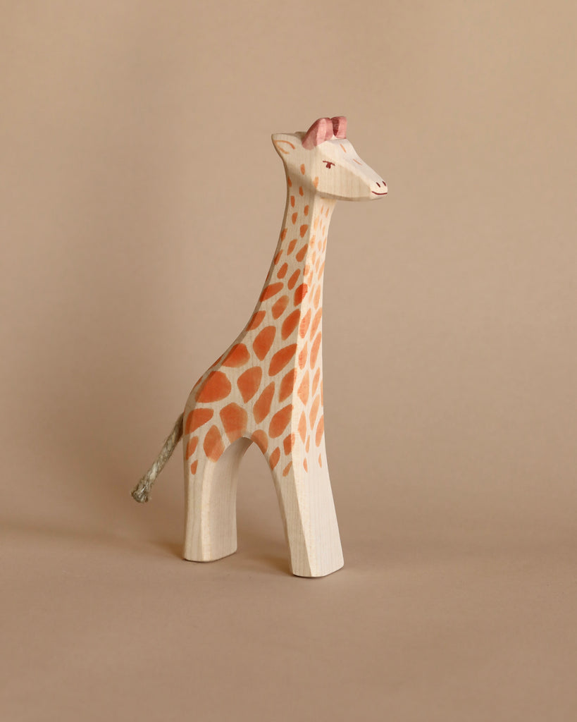 The Ostheimer Handmade Wooden Safari Animals Set features a handcrafted wooden giraffe with orange spots and a pink detail on its head, standing against a beige background—perfect for imaginative play.