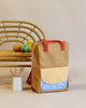 A beige Sticky Lemon Backpack Large with a blue and white checkered front pocket, crafted from waterproof nylon, stands next to a bamboo chair with colorful fruit and a cup of pencils on a neutral background.