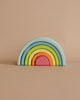 A Grimm's Small Wooden Rainbow, Pastel with six pastel arches. The largest arch is light blue, followed by green, yellow, orange, pink, and a small red arch at the center. This versatile toy is set against a beige background with no other objects in the scene.